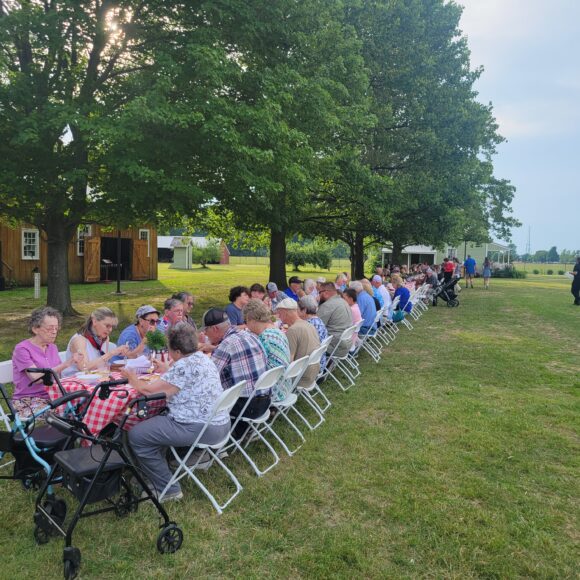 Long table in field with people eating a meal.