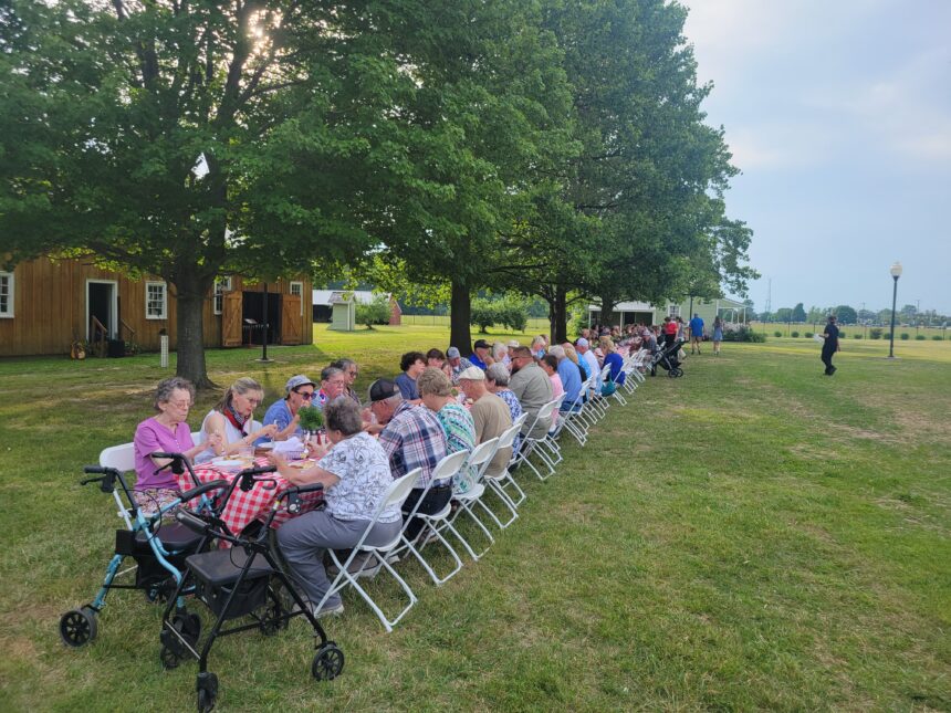 Long table in field with people eating a meal.